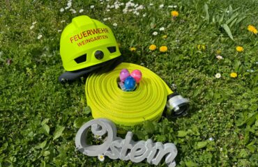 Es sind Ostereier zu sehen, die in einem aufgrollten Schlauch liegen. Links oben ist ein Helm auf den Schlauch gelegt, unten lehnt das Wort Ostern am Schlauch.